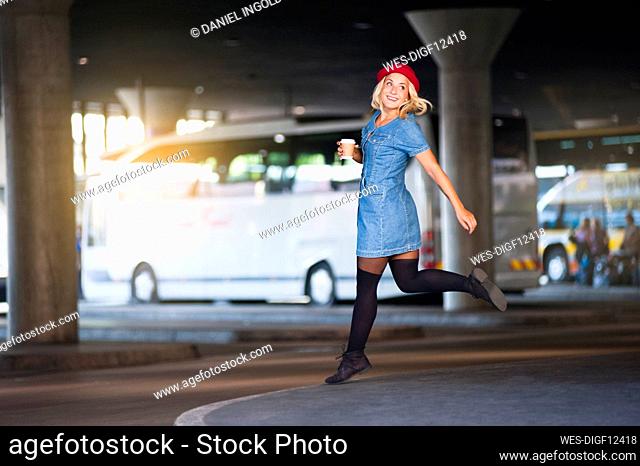Portrait of happy young woman with coffee to go jumping in the air at bus terminal