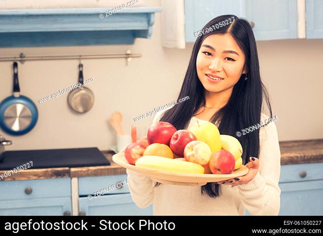 Toned picture of Chinese or Korean lady posing with bowl full of different fruits. Beautiful woman smiling for camera and showing fruits to camera at home