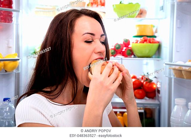 Smiling Young Woman Looking At Burger Standing In Front Of Refrigerator