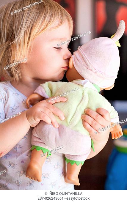 lovely and tender scene of blonde caucasian cute baby two years old kissing on the cheek or talking to ear a doll in hands indoor