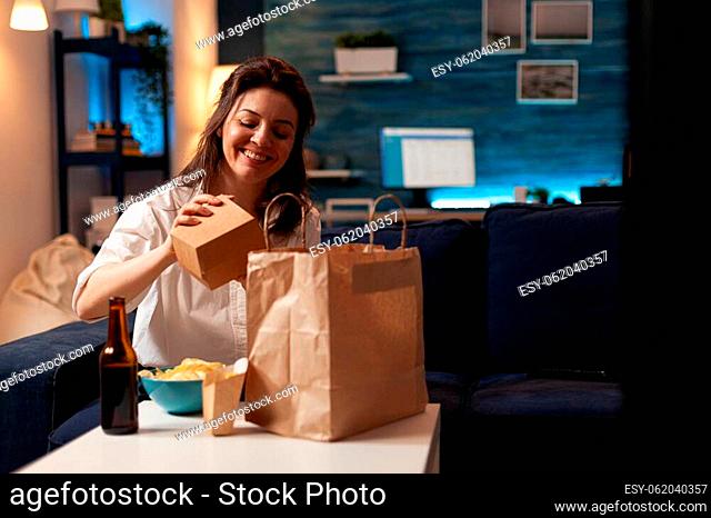Smiling cheerful woman unpacking tasting fast-food home delivered sitting on couch having lunch serivce order. Caucasian female enjoying delicious takeaway...