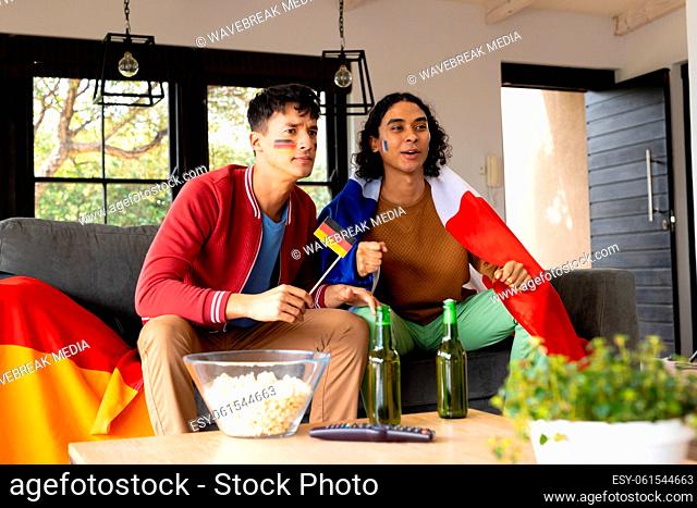 Happy biracial male couple sitting on sofa, supporting and holding flags of germany and france