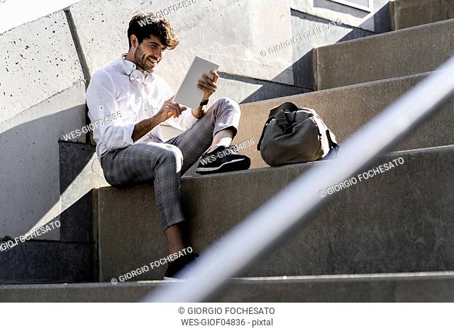 Smiling young man sitting on stairs outdoors using tablet