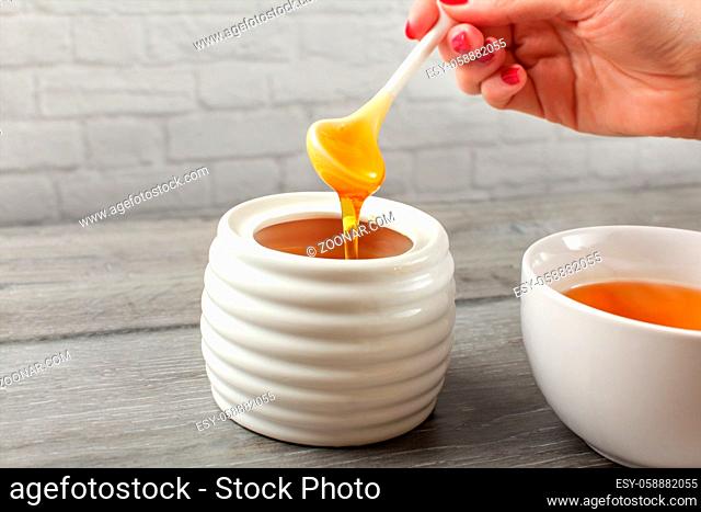Detail on woman holding small ceramic spoon, filled with honey, about to put it in white tea cup full of hot tea