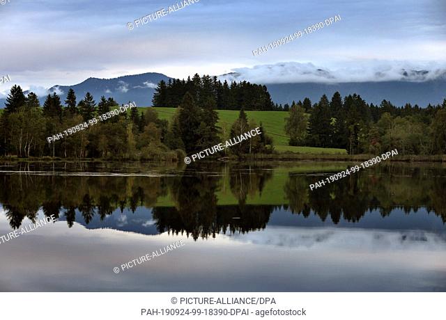 24 September 2019, Bavaria: The cloudy sky, the panorama of the Alps and trees are reflected shortly before sunset on the calm water of the Schmuttersee