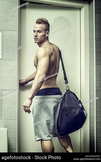 Shirtless muscular young man opening door, with gym bag on shoulder, shot from behind