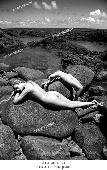 Two young adult nude Caucasian women lying on boulders on rocky coast