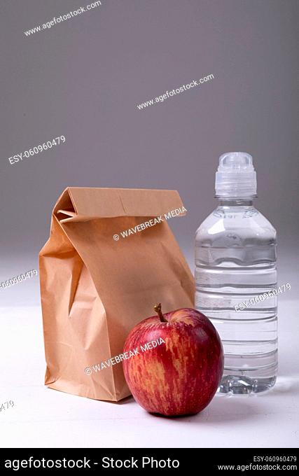 Close-up of brown paper lunch bag with apple and water bottle on table against gray background