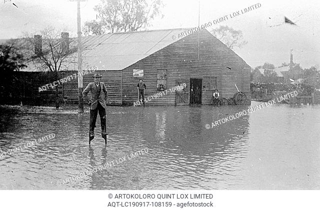 Negative - Rochester, Victoria, pre 1927, Rochester streets under floodwater. Two men on stilts stand in the floodwater in front of W. G