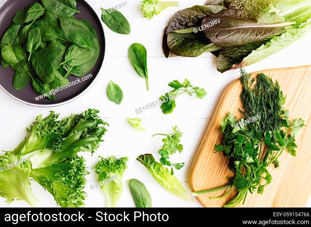 Flat lay with cutting board and bowl of fresh green salad leaves of spinach and lettuce, romaine and parsley, basil on white background