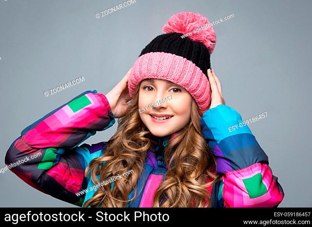 Beautiful happy teen girl with long curly hair in pink wool hat and bright warm coat. Studio shot over grey background. Copy space