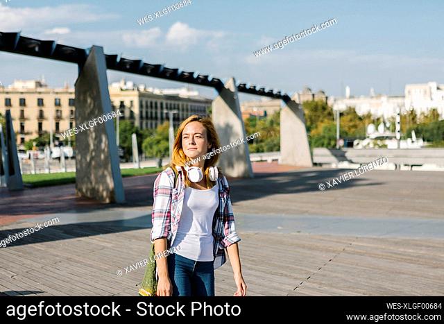 Young woman looking away with headphone around her neck walking on footpath during sunny day