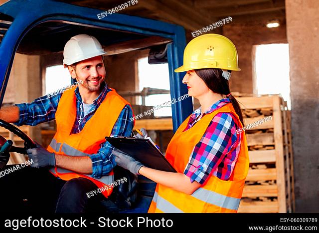 Male forklift driver sitting in a truck, female factory worker standing near, both discussing working issues. Warehouse workers in hardhats and orange...