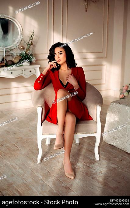 Pretty and sexy brunette woman in red dress sitting on luxury chair, posing at camera and leaning by handr. Seductive young girl with wave hair
