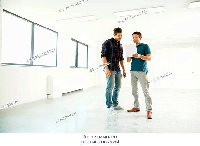 Two men standing in empty office space, looking at digital tablet