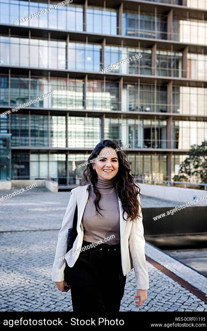 Smiling young businesswoman walking in front of modern office building