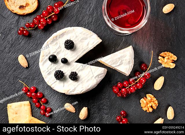 An overhead photo of Camembert cheese with a glass of red wine, fruits and nuts, shot from above on a black background