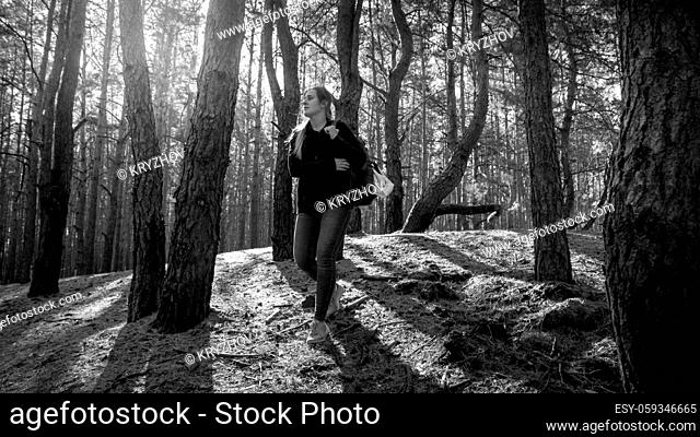 Black and white photo of beautiful young woman with backpack hiking in the old forest