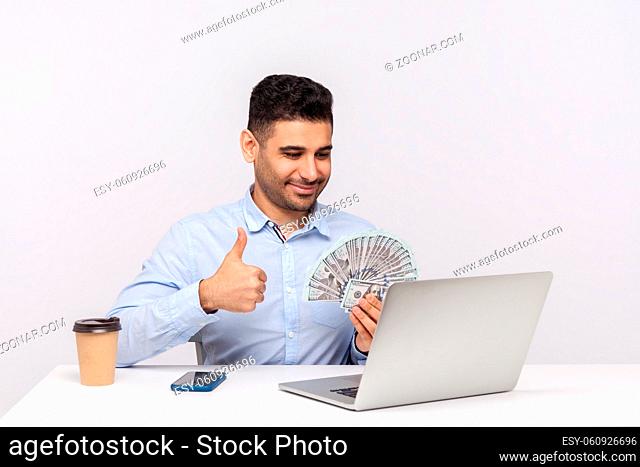 Successful happy businessman sitting in office workplace holding money dollars and showing thumbs up to laptop screen, having online conversation