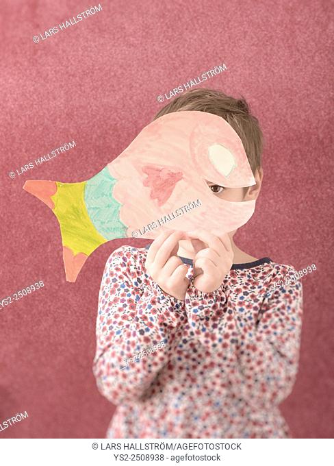Portrait of little girl in dress holding a cut out drawing of a fish in front of her face. She is shy and hiding behind it