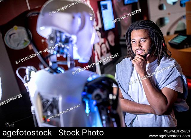 Thoughtful technician with hand on chin looking at human robot in workshop