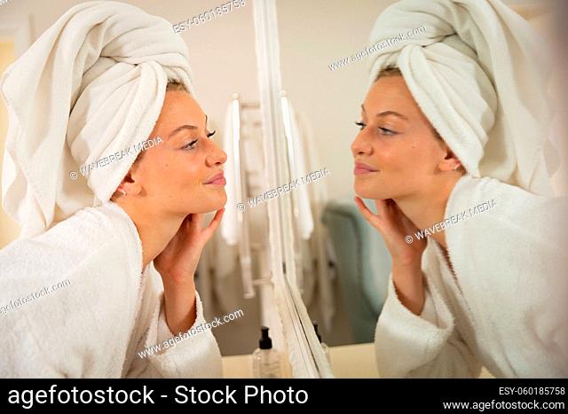 Smiling caucasian woman in bathroom with towel on head, looking in mirror and moisturising face
