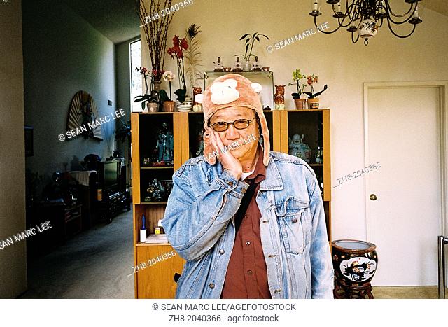 An older Asian man wears a monkey hat while holding his hand on his cheeks in the living room of his Californian home