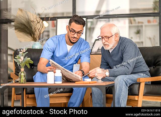 Focused old man seated at the coffee table with his caregiver reading an article in the newspaper