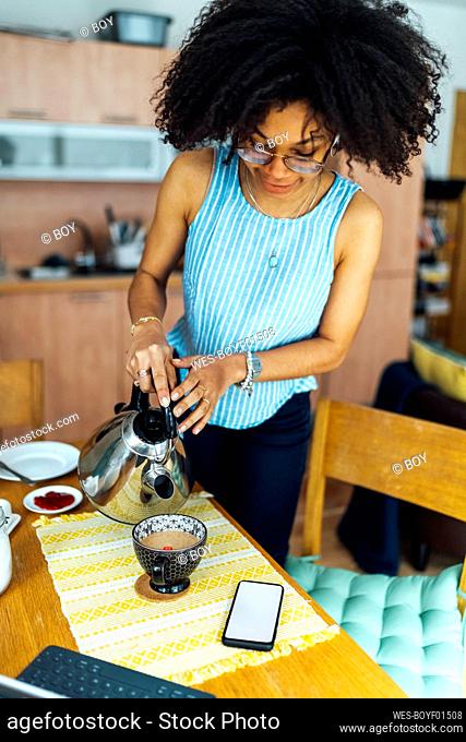 Young woman with curly hair pouring water in coffee cup on table at home