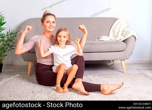 Attractive fit mom and her charming daughter showing biceps, smiling at camera, sitting together on floor after yoga workouts at home