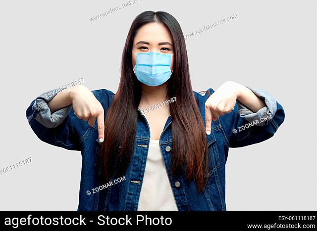 Here and right now. Portrait of serious bossy brunette asian young woman with surgical medical mask in blue denim jacket standing and pointing down