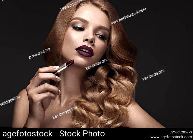 Beautiful blonde in a Hollywood manner with curls, dark lips, lipstick in hand. Beauty face and hair. Picture taken in the studio
