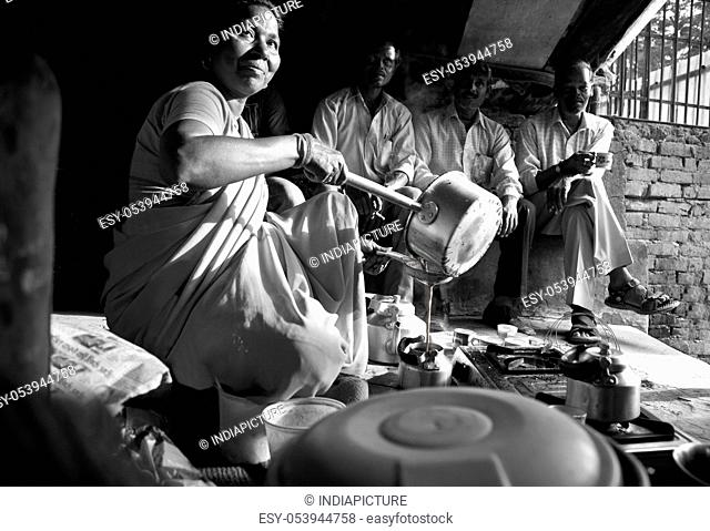 Black and white image of Indian female vendor pouring tea in kettle with customers sitting in background