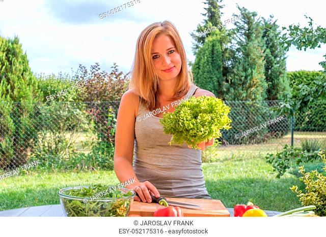 Attractive young woman is preparing vegetable food outdoors. Woman is offering a head of green leaf lettuce looking at the camera