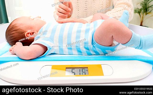 Young caring mother stroking her little newborn baby son while weighing him on digital scales in hospital. Concept of babies and newborn hygiene and healthcare