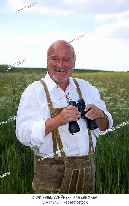 Elderly man wearing a traditional pair of trousers and holding binoculars in his hands in a corn field