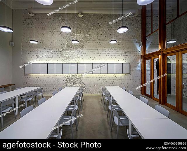 Luminous hall in the cafe with shabby light walls and gray floor. There are white tables with chairs, frames on the wall, hanging round lamps