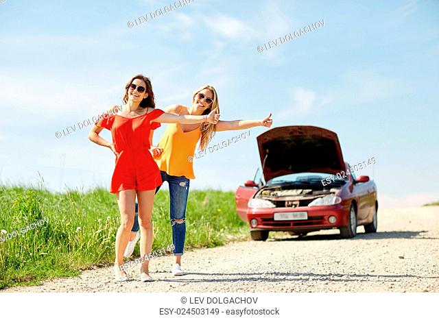 road trip, transport, travel, gesture and people concept - happy young women with broken car showing hitchhiking gesture asking for help at countryside