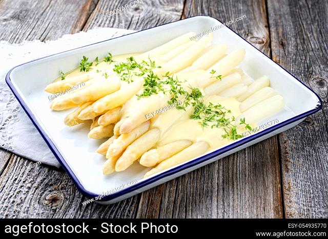 Traditional steamed white asparagus with herbs as closeup in an enamel tray on a wooden board