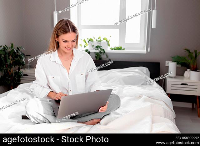 Pretty smiling Caucasian girl sits on bed in white-gray bedroom and use laptop for remote work, talking on video call. Communication, technology