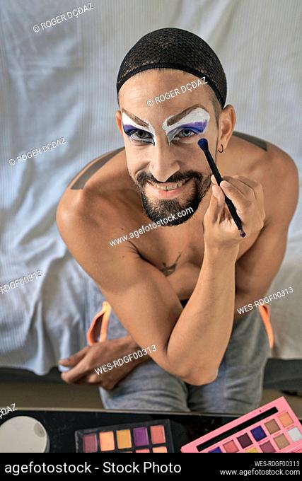 Smiling shirtless man with blue eyeshadow on eyes holding make-up brush