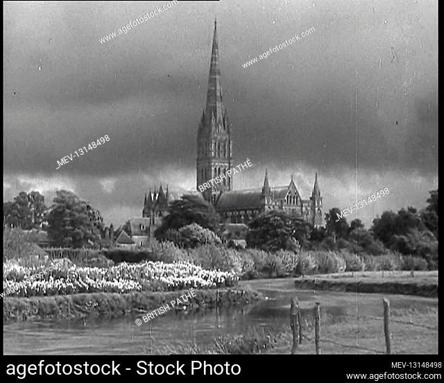 A Pond and Flowers in Bloom on a Bank Alongside and Mature Bushes and Trees Beyond That in Front of a Large Church With a Tall Spire Under a Cloudy Sky - United...