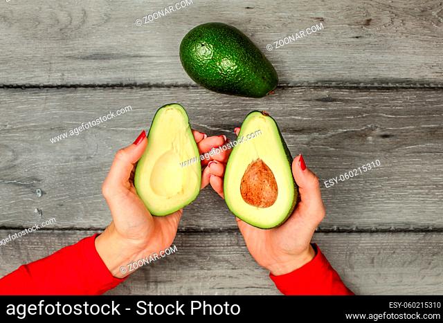Tabletop view woman's hand holding two avocado halves, seed visible, whole avocado pear next to it