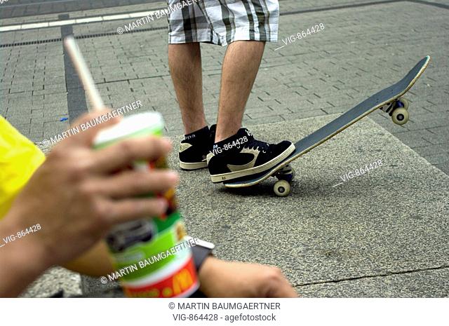 skateboarding. Our picture shows in the foreground the hand of a young mit with a drink in a plastic cup, and behind him an other one skateboarding