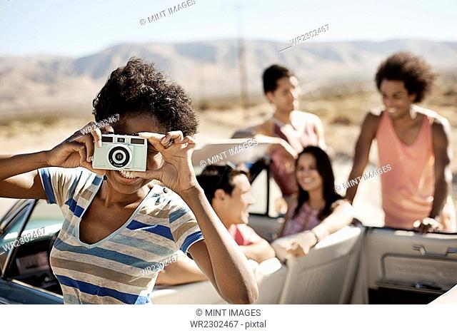 A group of friends by a pale blue convertible on the open road, on a flat plain surrounded by mountains, one holding a camera