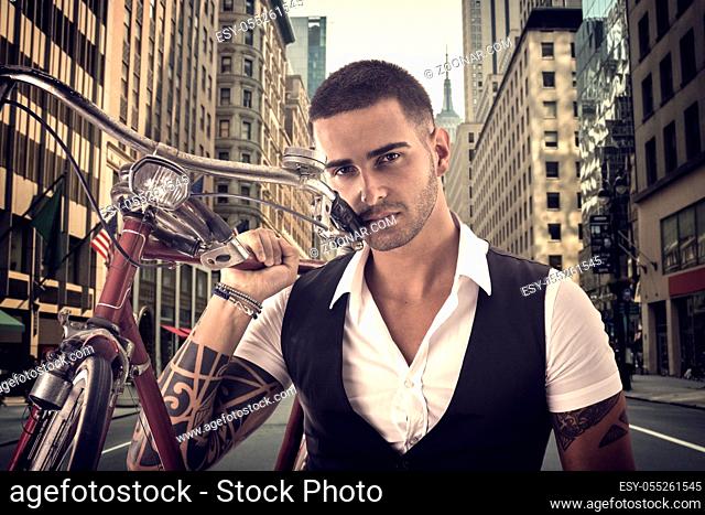 Portrait of young tattooed man in elegant clothes holding a bicycle in middle of a city street