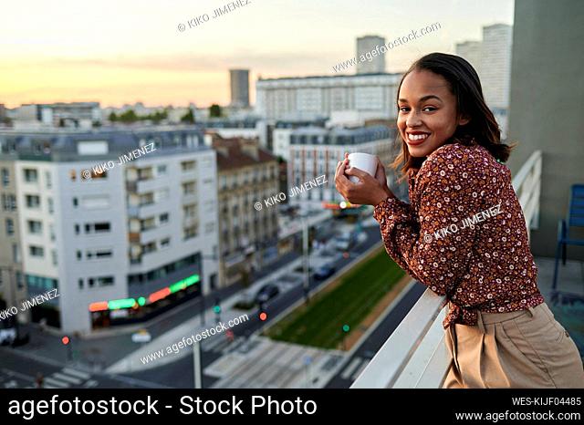 Smiling young woman with coffee cup in balcony at sunset