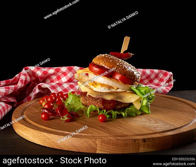 big delicious burger with meat cutlet, cheese, fried egg, tomatoes, cucumber slices and green lettuce, fast food on a round wooden board, black background