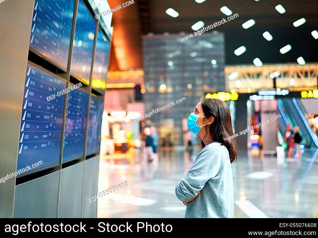Woman in virus protection face mask looking at information board checking her flight in international airport. Departure board, flight status
