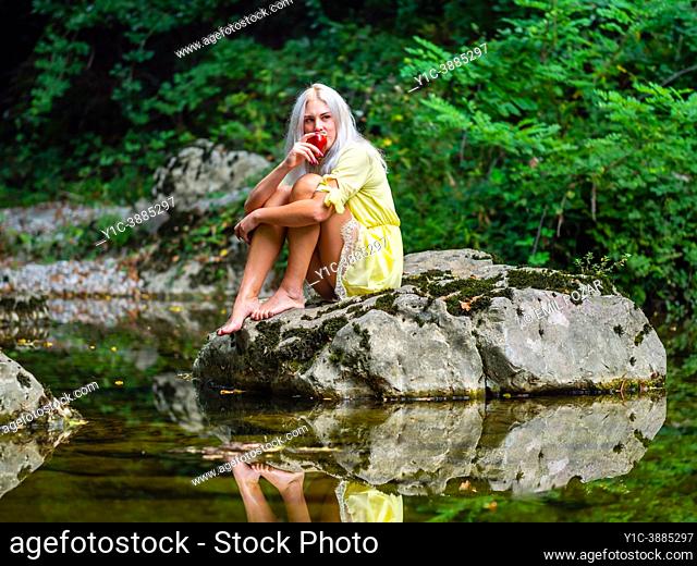 Attractive blonde woman on a Green forest river eating Red apple